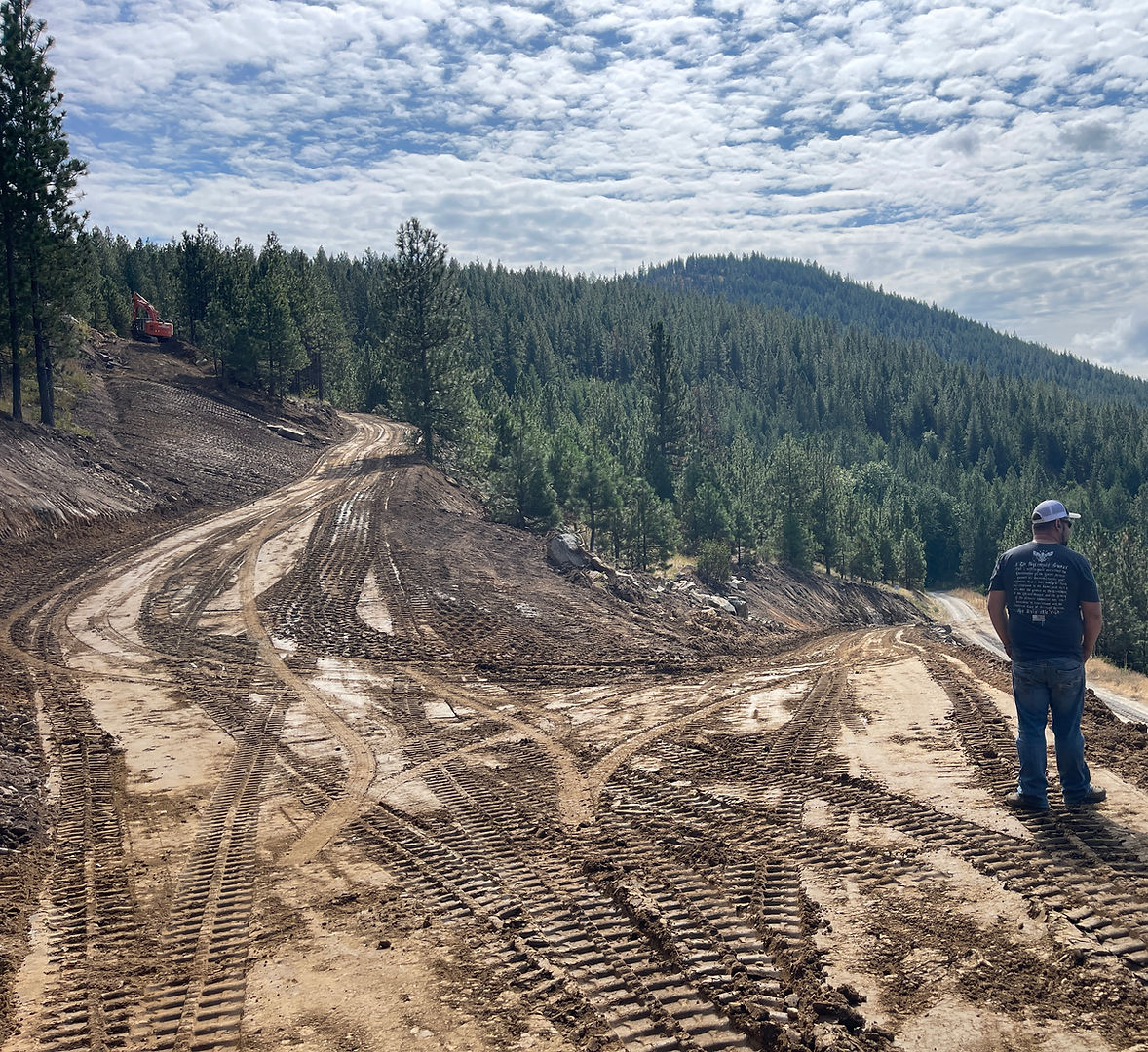 Contractor surveying a freshly graded hillside road cut through pine forest with an excavator working in the background in North Idaho<br />
