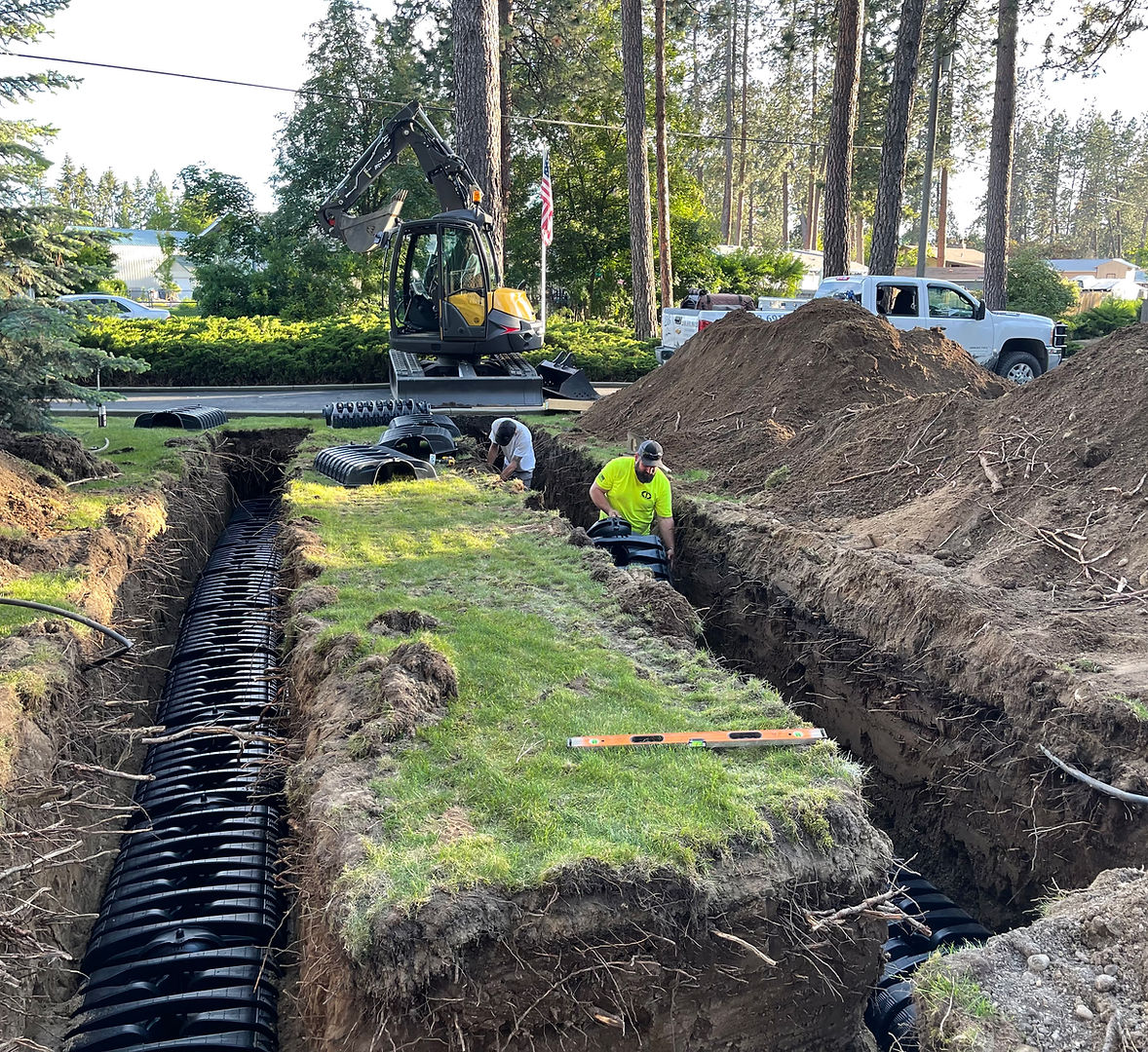 Two workers installing a septic drain field system with a yellow excavator and black leach chambers visible in open trenches on a residential property in North Idaho<br />
