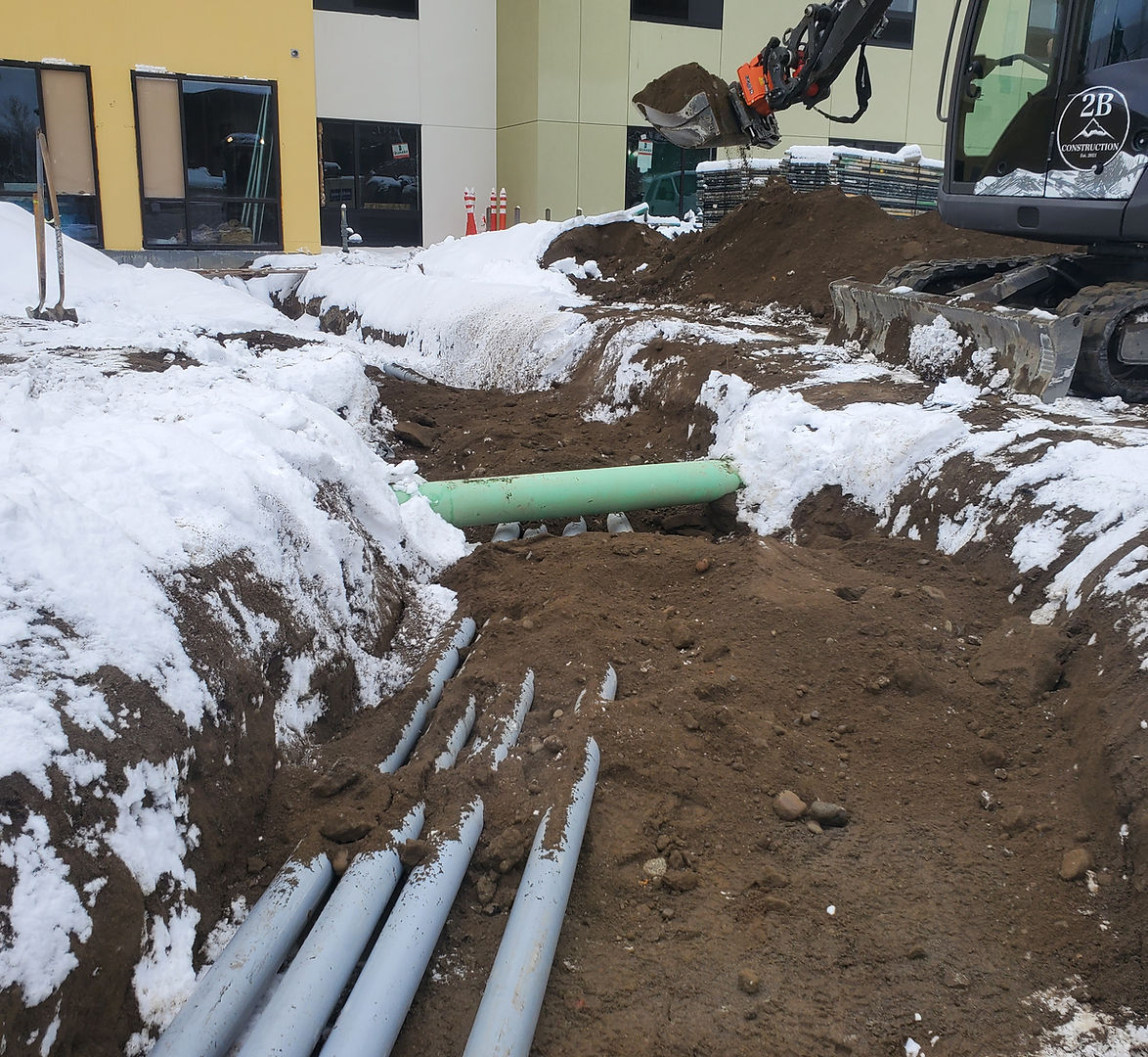 Excavator trenching next to a commercial building in winter with multiple utility pipes being laid in a snow-covered job site<br />
